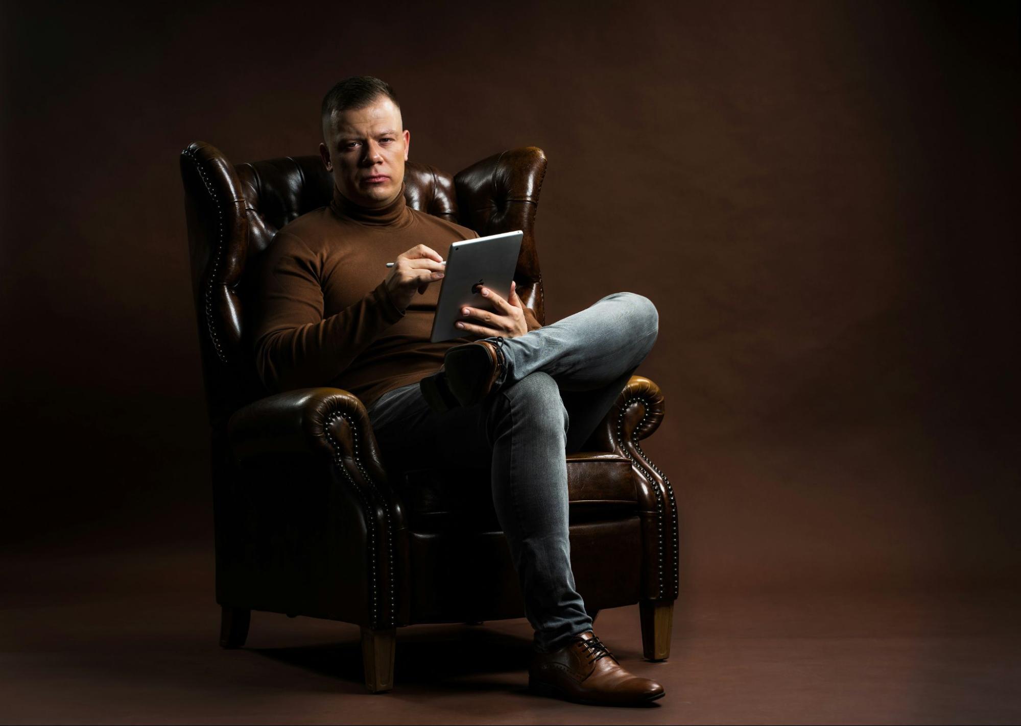 Un homme trône sur un fauteuil jambes croisées avec une tablette à la main.